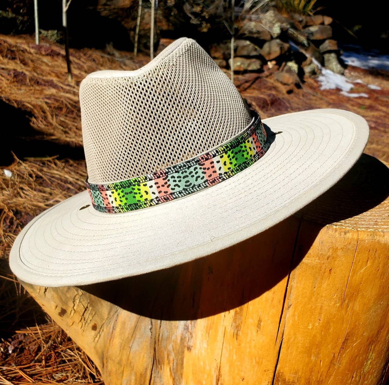 Light beige river hat leaning on wooden stump, featuring rainbow trout patterned hat band. Hat band has yellow, green, pink, blue coloring with black speckles mimicking the coloring of rainbow trout.