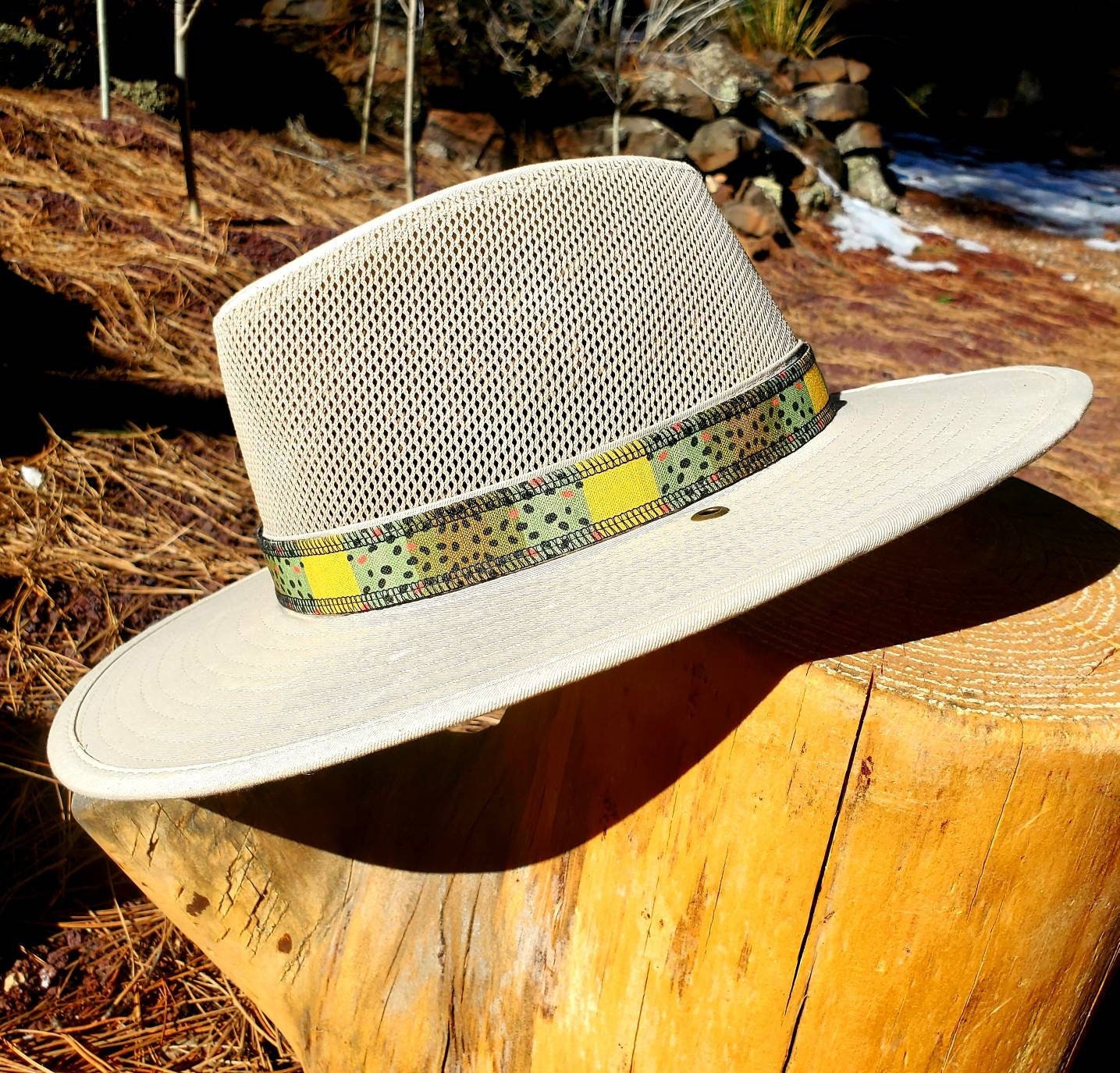 Light beige river hat leaning on wooden stump, featuring brown trout patterned hat band. Hat band has yellow, green, brown coloring with black speckles mimicking the coloring of brown trout.