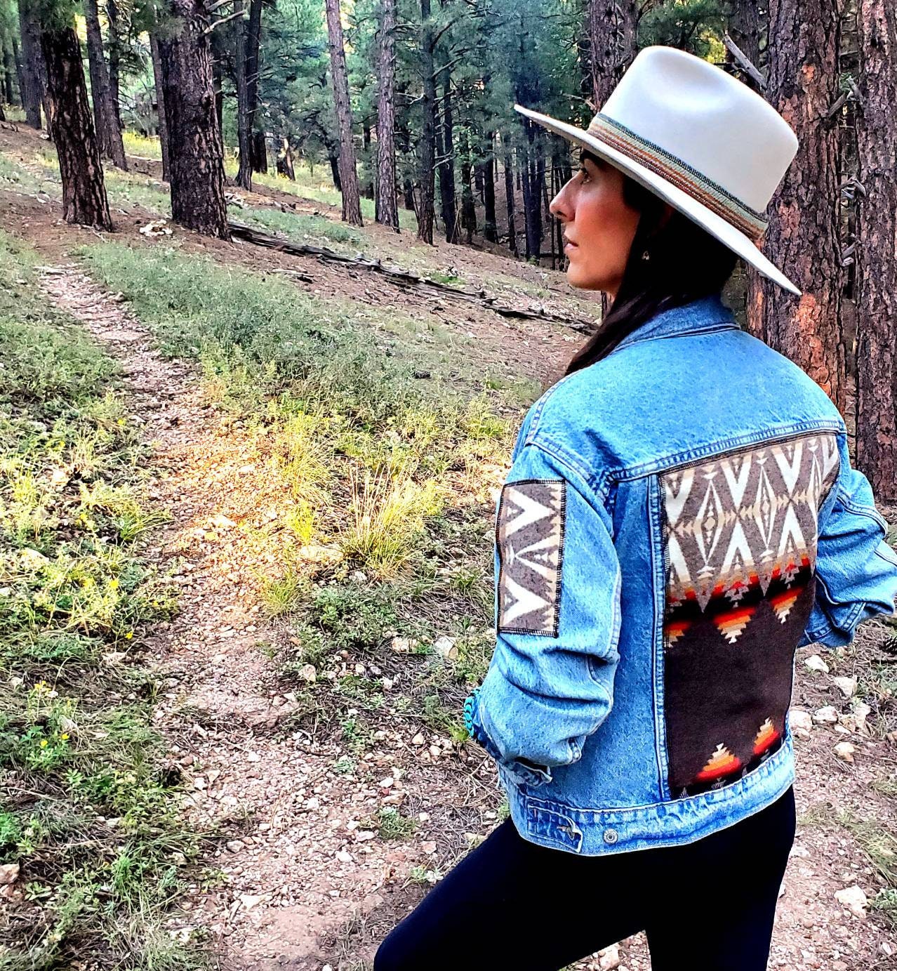 Model is standing in the woods wearing upcycled denim jacket with Pendleton® wool patches on sleeve and back panel.