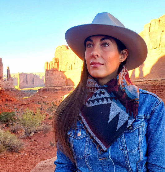 Model is at sitting amongst red rock outcroppings wearing a wool wild rag, cowl scarf for women, featuring southwestern, aztec designs.