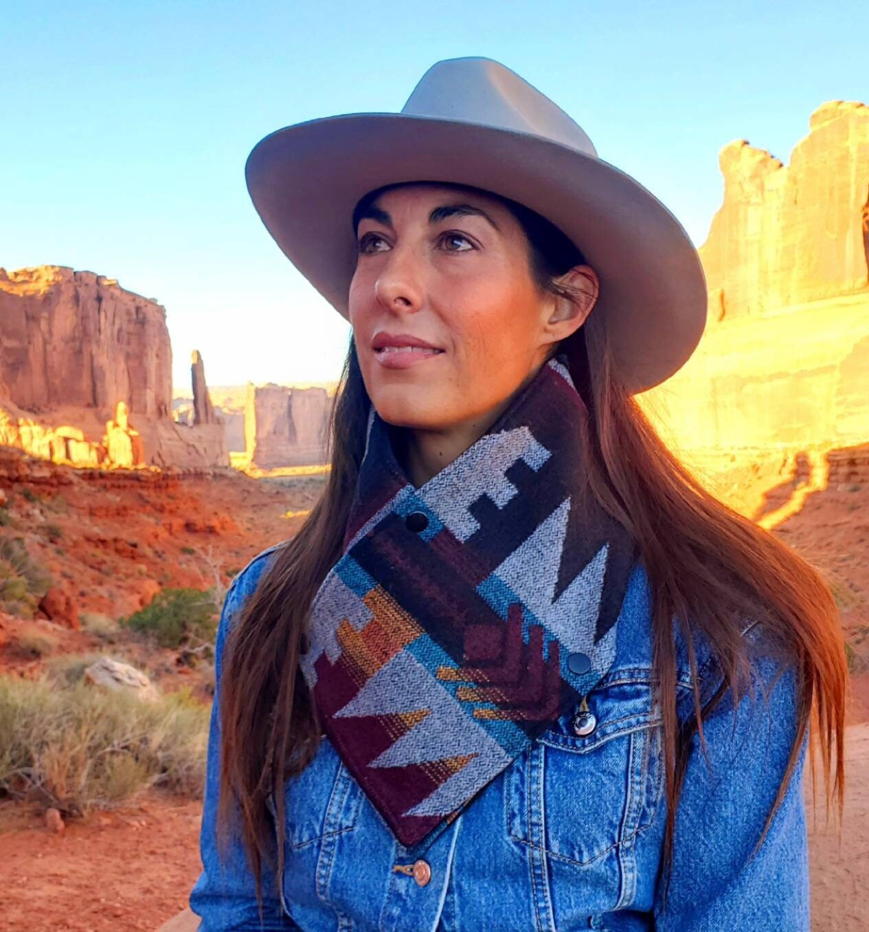 Model is sitting below Park Avenue at Arches National Park, wearing a southwestern wool cowl scarf, featuring burgundy, grey, black and turquoise geometric shapes that are common in the Southwest.