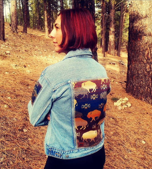 Model is standing in woods wearing southwestern denim jacket with Pendleton® wool buffalo fabric sewn into back panel and arm patches.