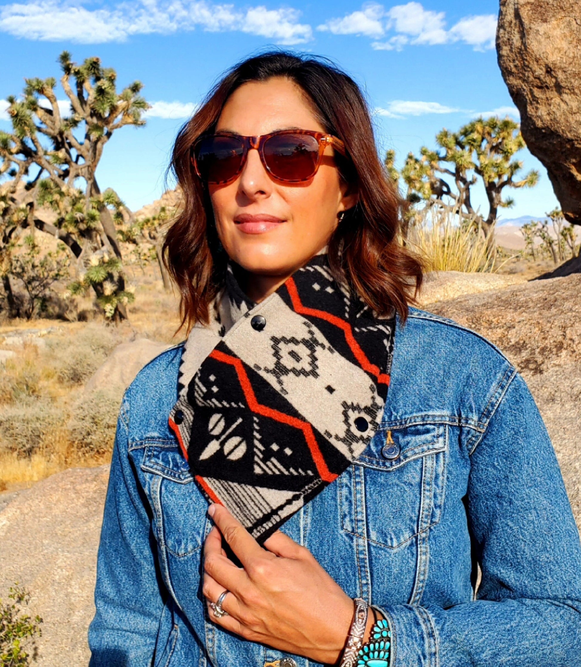 Model standing in front of Joshua Tree, wearing western cowl, made from wool & lined with fleece. Wool Wild Rag is black, tan and orange & features southwestern designs.