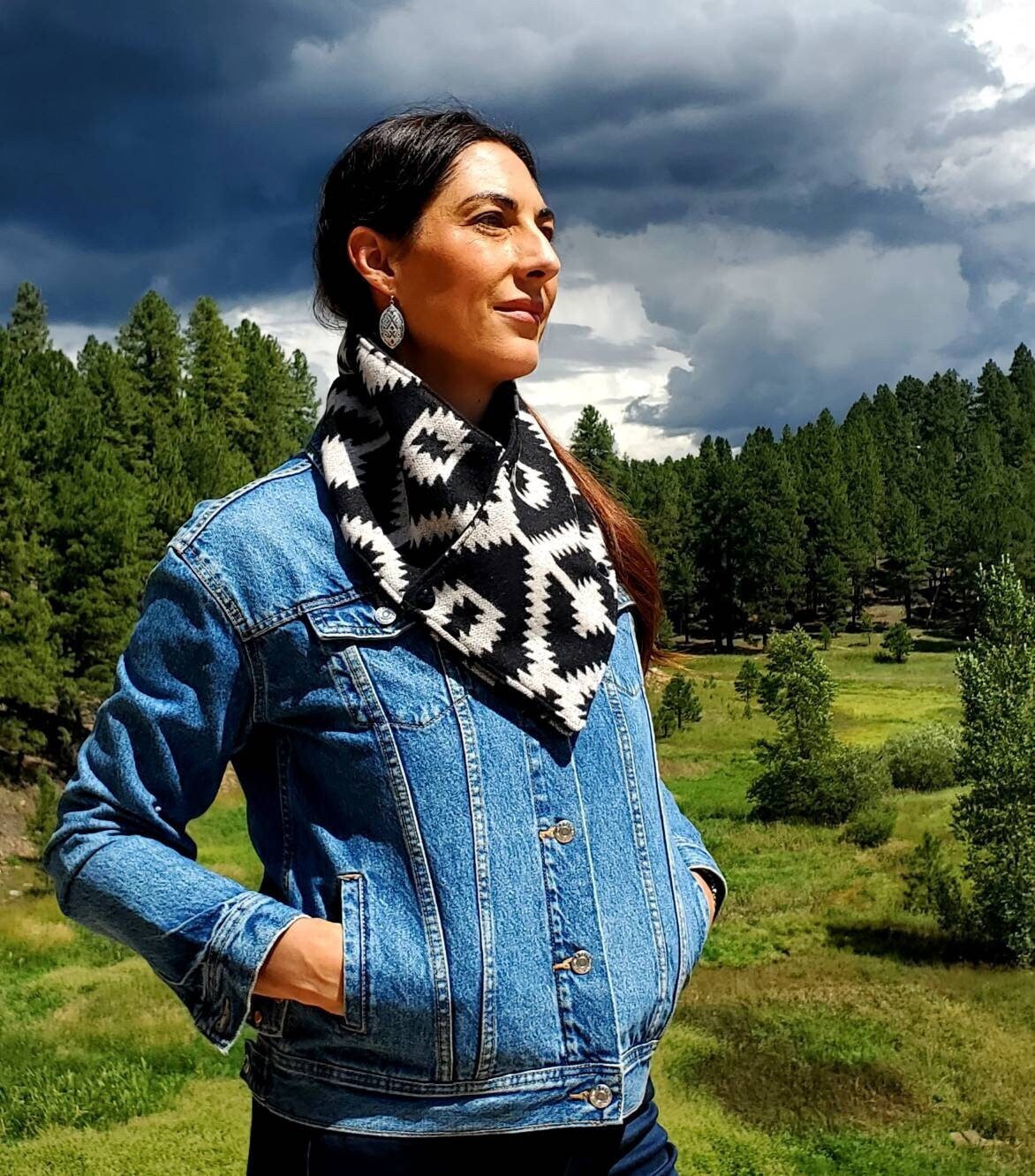 Model is at standing in forest wearing a black and white wool wild rag, cowl scarf for women, featuring southwestern, aztec designs.