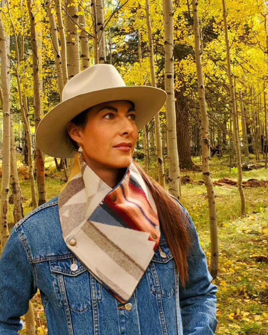 Western cowl neck scarf is worn by model standing amongst aspen trees. Looks similar to bandana or neck gaiter. Made with Pendleton® blanket-weight wool, the design features pink, brown, tan, blue geometric shapes that are popular in the Southwest.