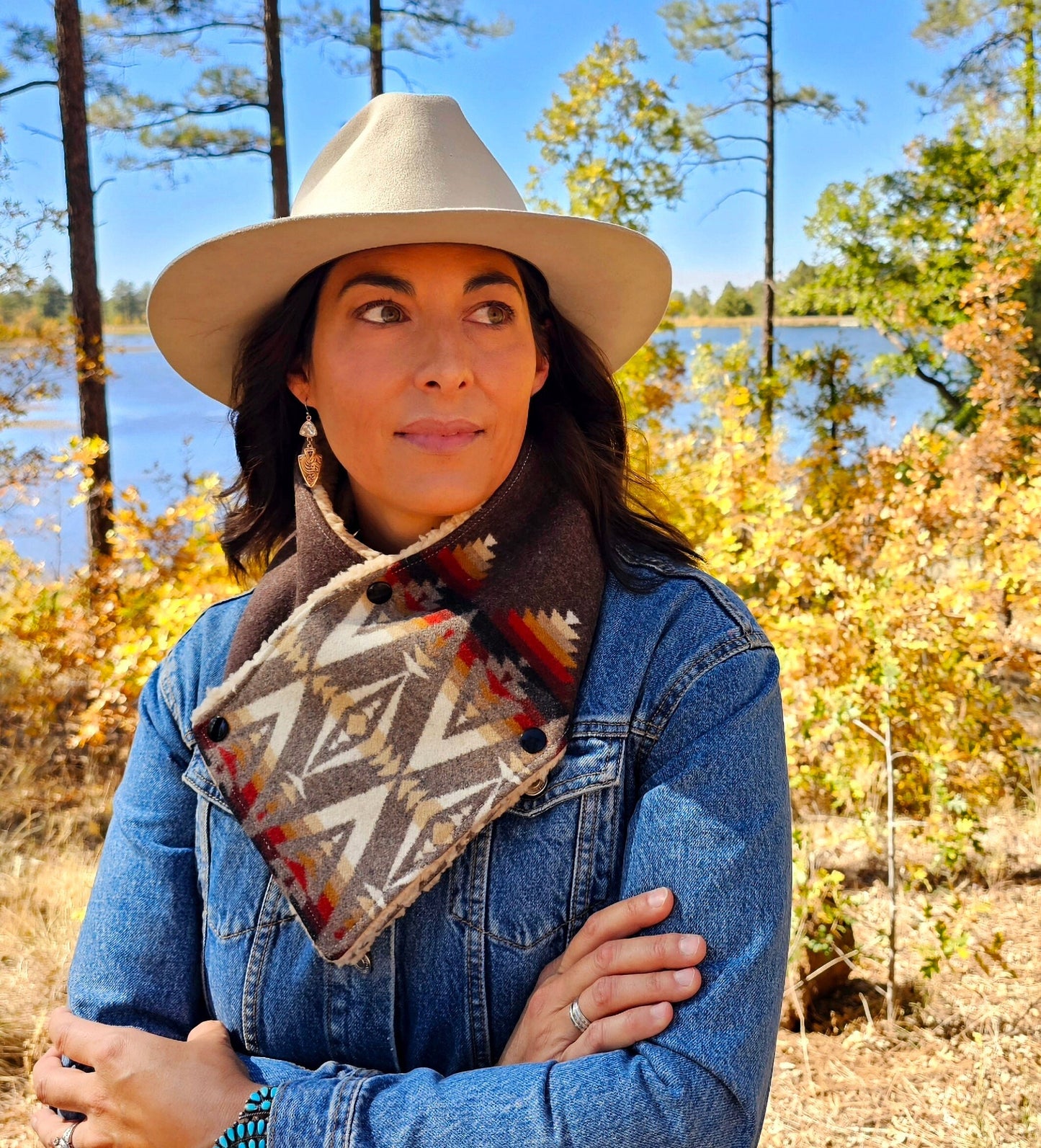 Model is standing in tree grove, wearing western cowl scarf. Bandana scarf is made from wool & sherpa, features red, white, black & brown geometric designs.