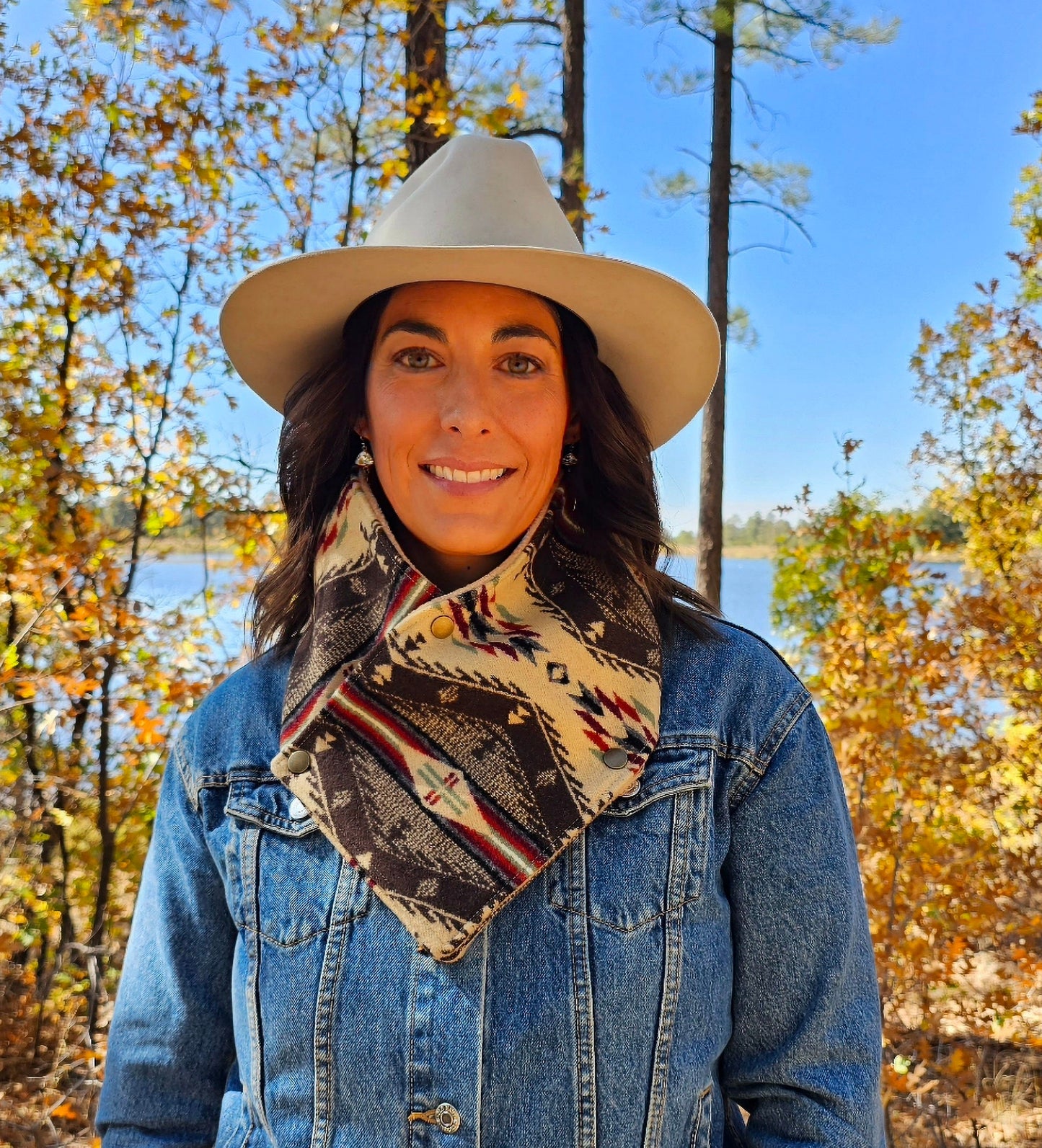 Model is standing in tree grove, wearing western cowl scarf. Bandana scarf is made from wool & cotton, features red cream, brown & seafoam geometric designs.